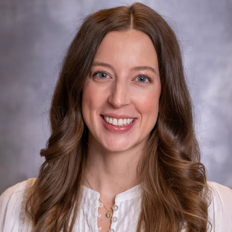 Professional headshot of a smiling woman with long, wavy brown hair and blue eyes, wearing a white blouse against a soft gray background.