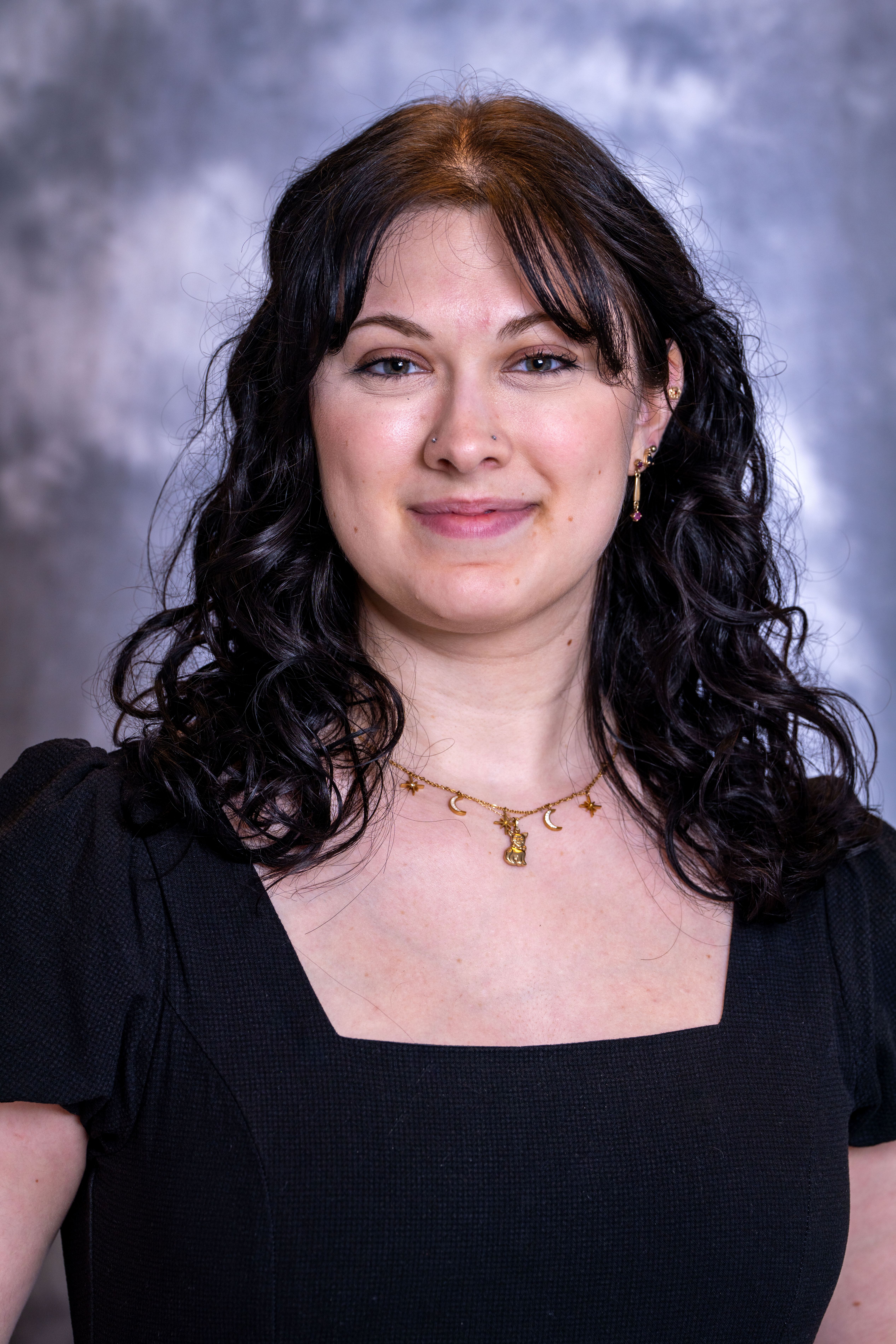 Young adult female with long black hair, curled at the ends. She is smiling and wearing a black dress. 