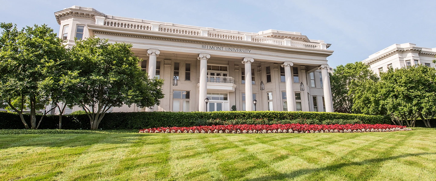A wide-angle view of Belmont University’s main building with tall white columns, manicured green lawn, and a vibrant flower bed in the foreground.