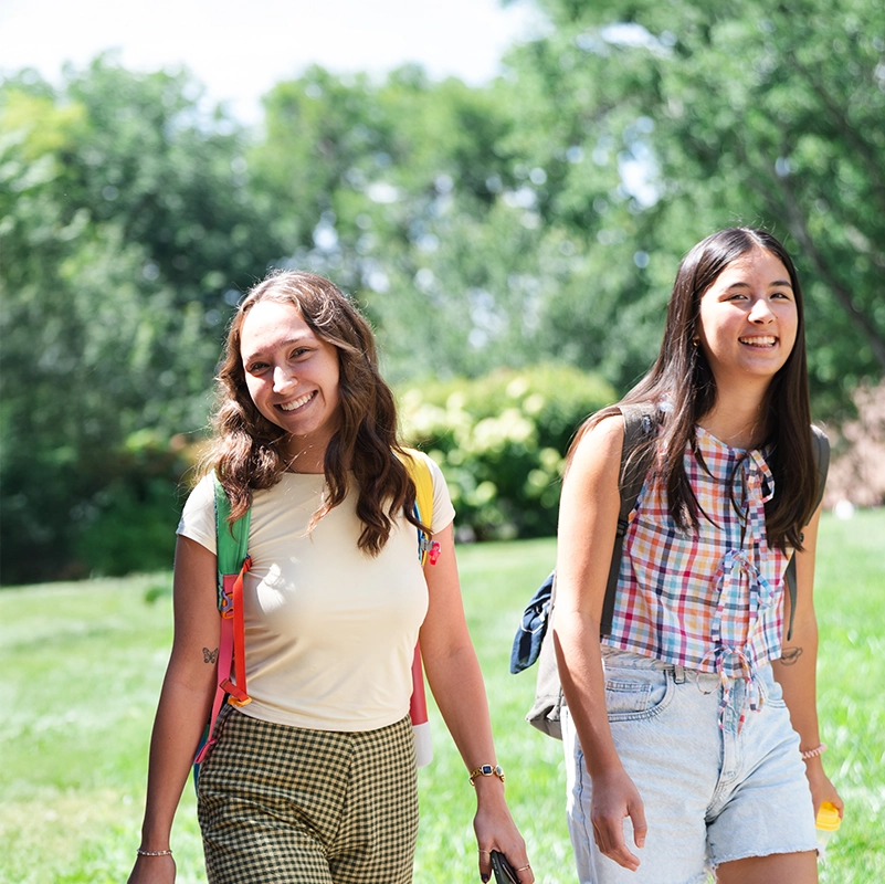 Two smiling students walk outdoors on a sunny day with backpacks on, surrounded by green grass and trees, enjoying a bright and welcoming campus atmosphere.