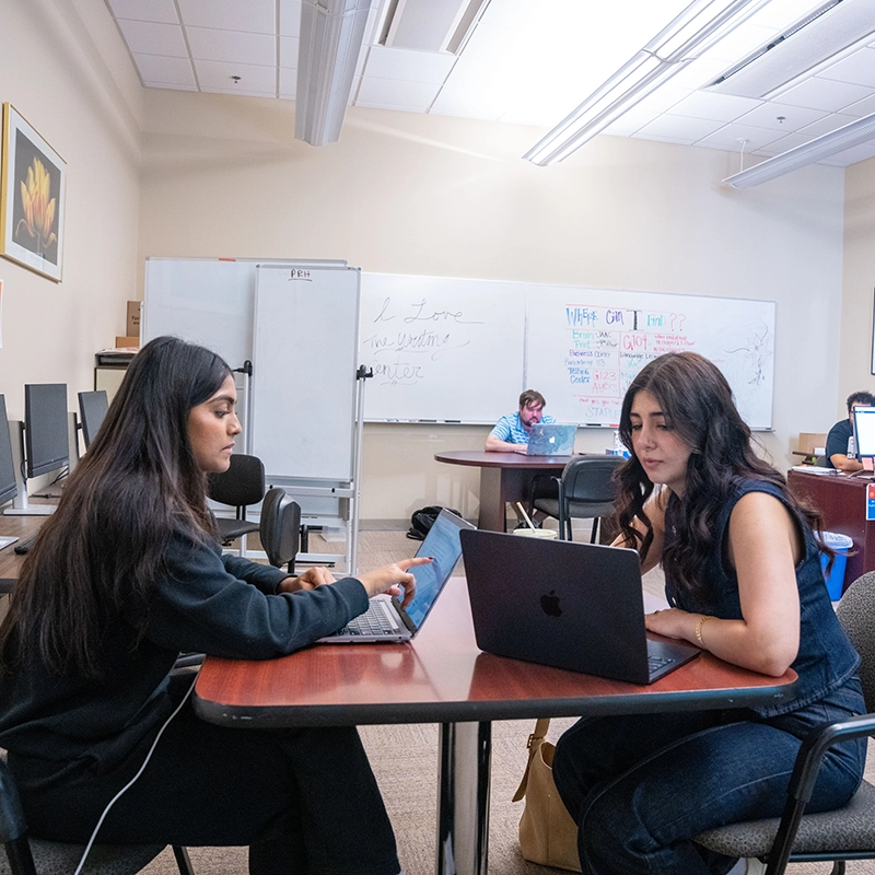 Two students work together on laptops at a table in a computer lab, with other students studying in the background and notes written on a whiteboard.