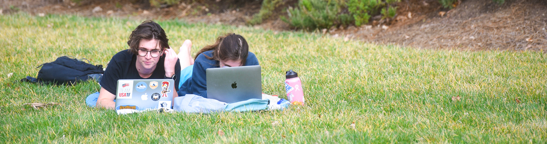 A photo of two Belmont students laying in the grass working on laptop computers.