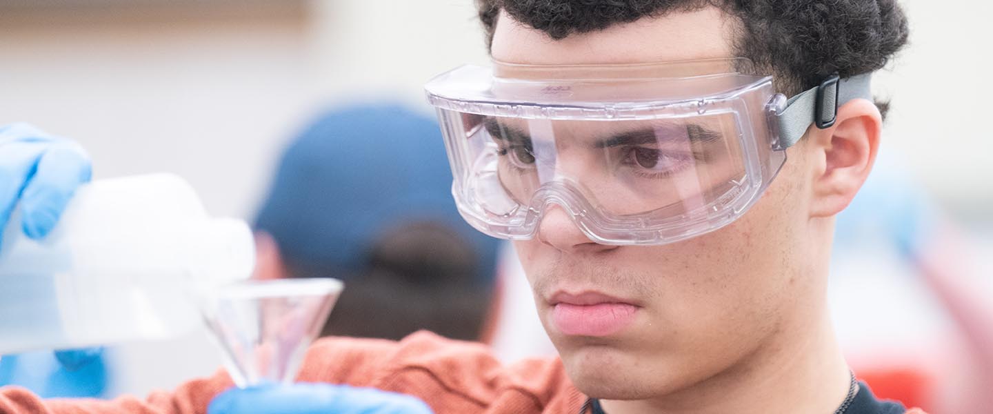 Student pouring liquid into beaker while wearing safety goggles