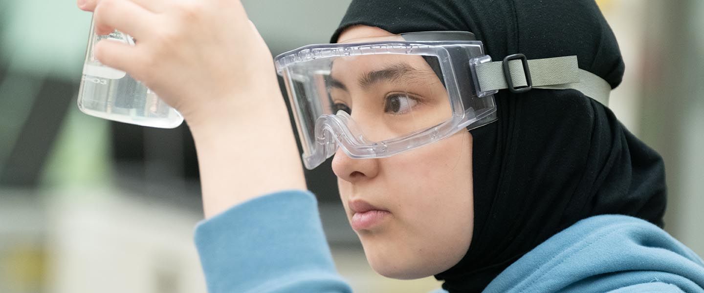 Student measuring liquid in beaker while wearing safety goggles in a classroom