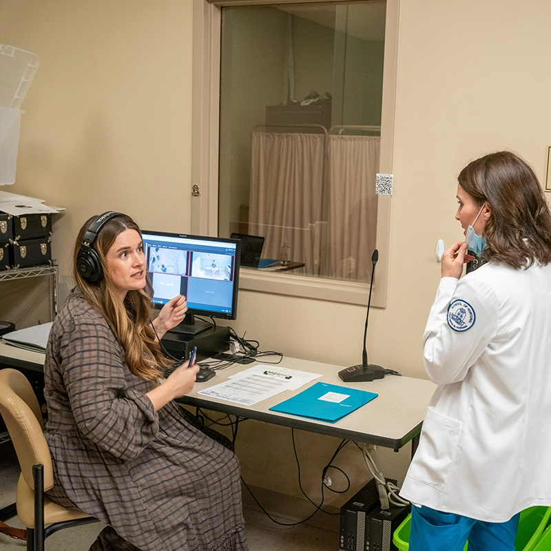 Nursing students converse in a simulation lab. One student is seated and looking at a computer while wearing headphones and is conversing with a standing nurse in a lab coat.