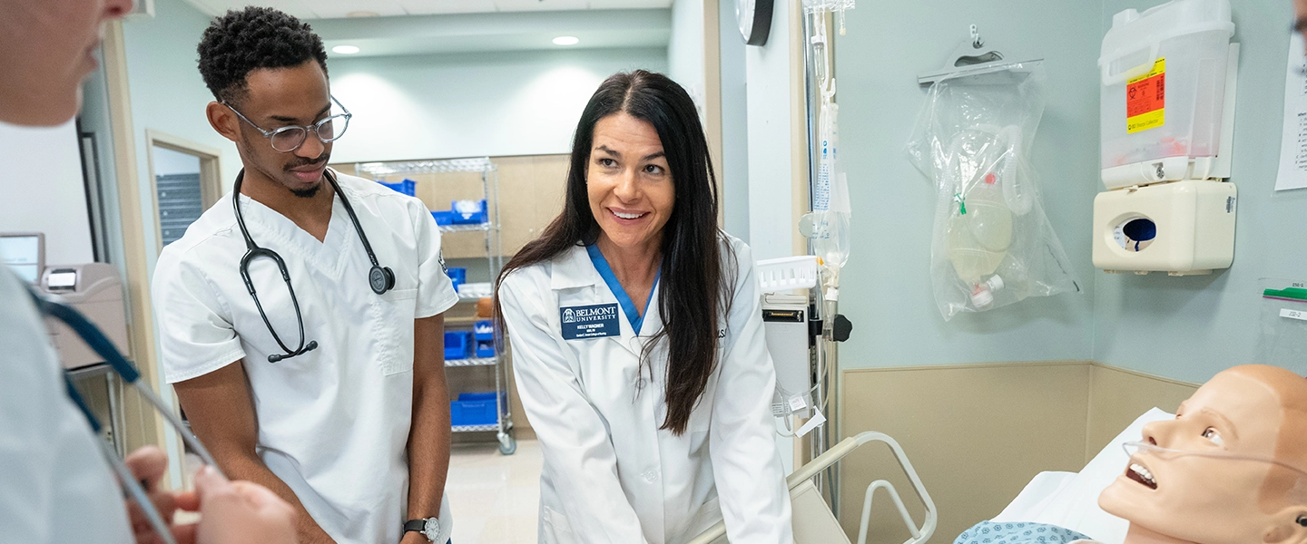 Belmont University healthcare instructor guiding nursing students during a simulation exercise with a medical mannequin in a clinical lab setting.
