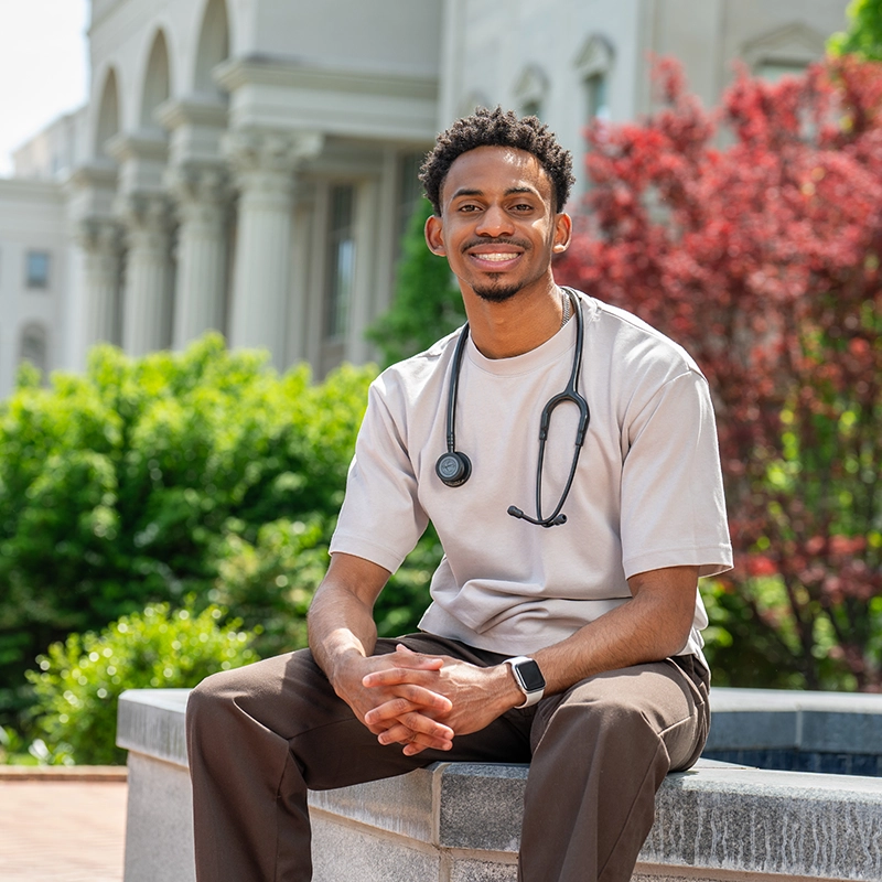 Belmont University pre-health student with a stethoscope sitting outside on campus, smiling confidently.