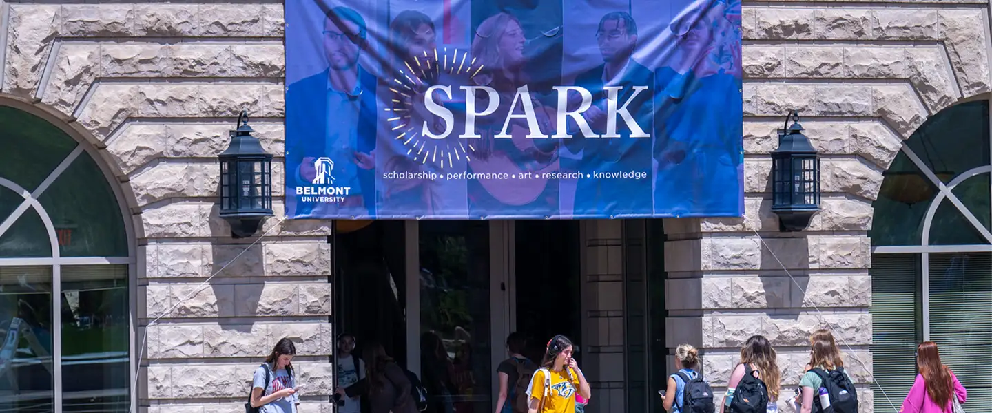 A large banner advertising "SPARK," Belmont University's student symposium, hangs over the entrance of a building. The banner features the tagline "scholarship • performance • art • research • knowledge." Students are seen walking in and out of the building, creating a lively atmosphere on campus.
