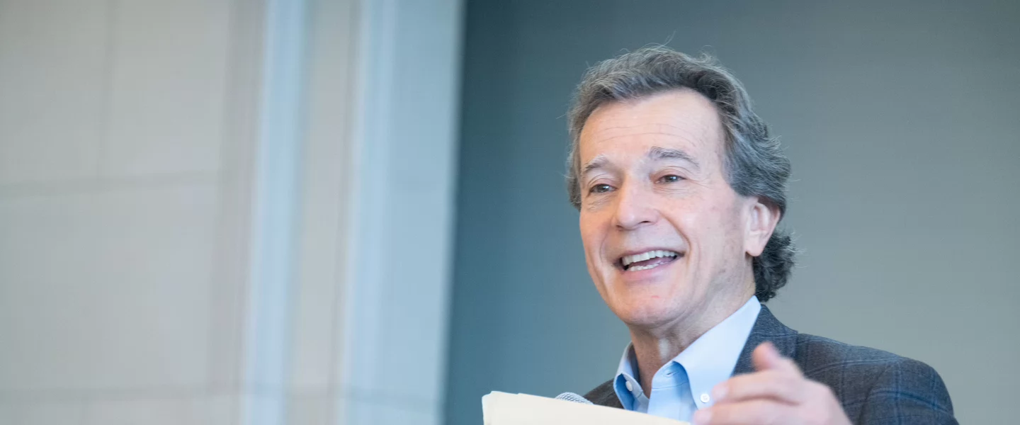 A man in a suit gestures energetically while holding papers, smiling as he speaks. The light background and his expression suggest a positive tone.