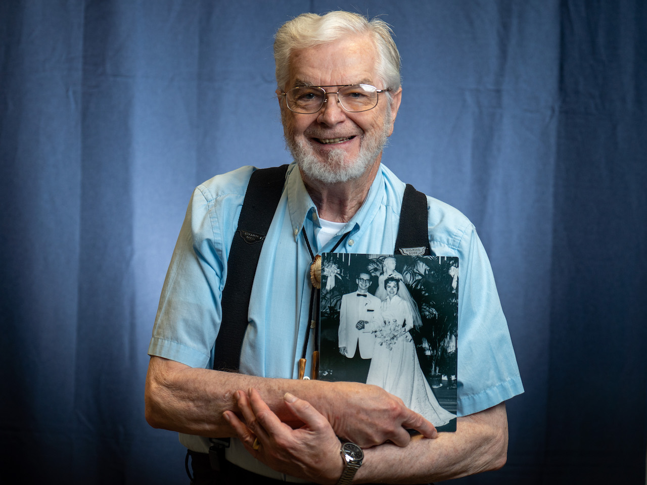 The image shows an elderly man with white hair and beard wearing a light blue button-up shirt with black suspenders against a blue backdrop. He's smiling while holding a black and white wedding photograph, presumably from his younger years. The wedding photo shows a couple - a man in a white jacket and bow tie alongside a bride in a traditional white wedding dress holding a bouquet. The man is wearing glasses and a wristwatch, and his expression suggests he's fondly reminiscing about this significant moment from his past.