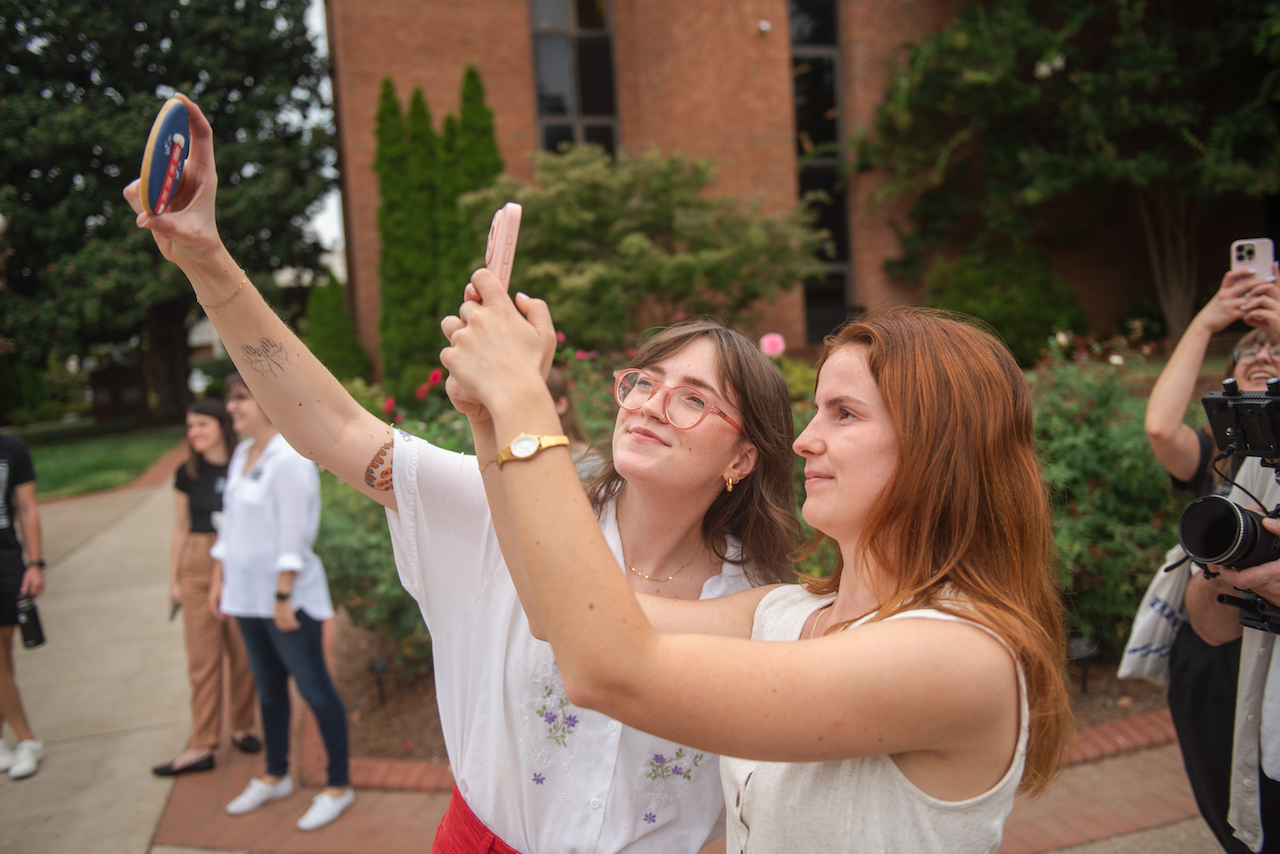 Two individuals outside on Belmont's campus. One person, wearing a white shirt with purple embroidery and red pants, is holding up a decorated Belmont-themed cookie while another person takes a photo with their phone.