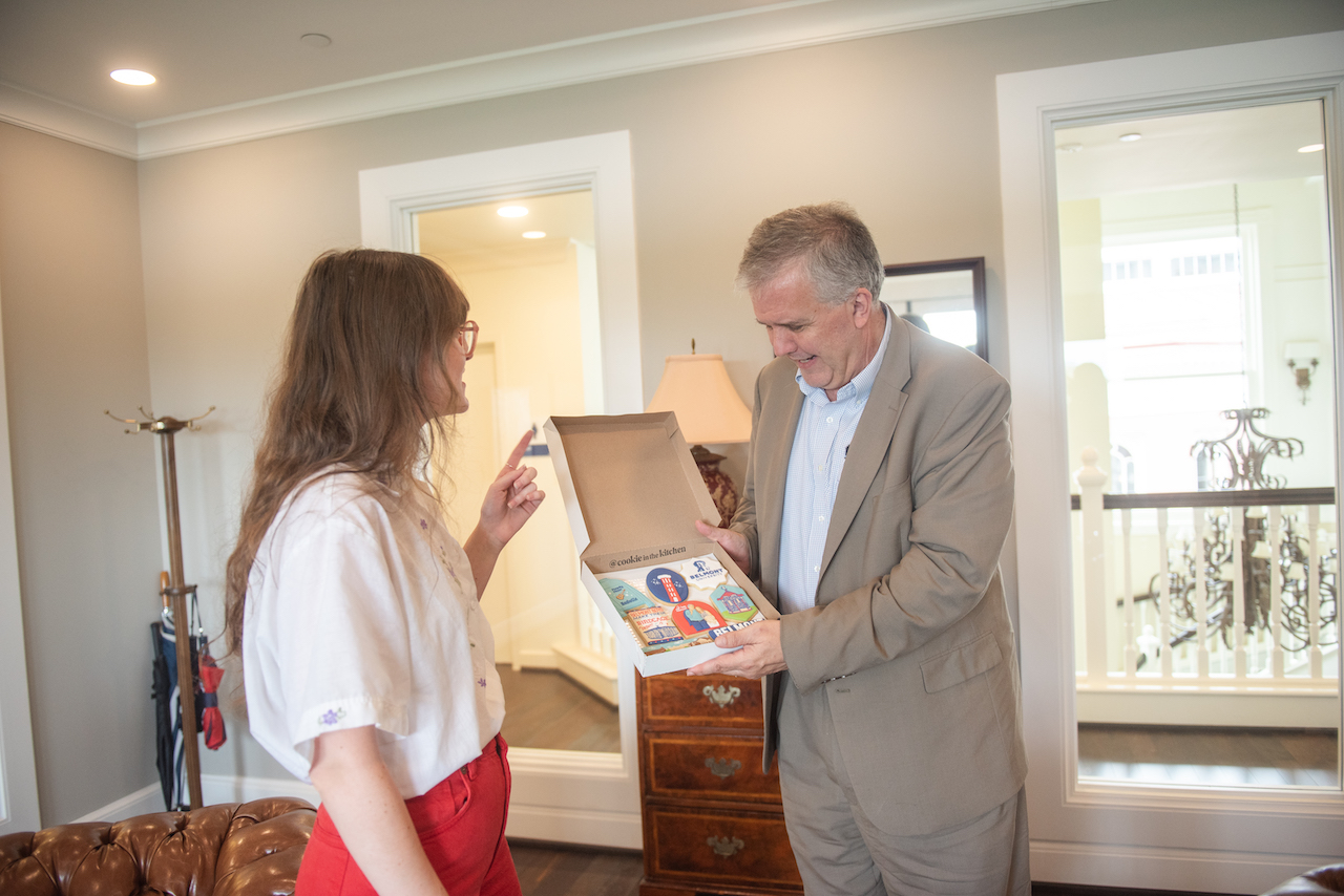 An indoor scene showing the same person in white shirt and red pants presenting the box of Belmont-themed cookies to an older man in a light gray suit, who is examining the cookies with interest.