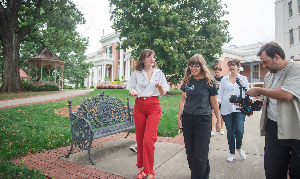 A group walking on Belmont's campus. The scene includes the person in white shirt and red pants talking with someone in a black shirt, followed by others including a cameraman, with historic university buildings and a gazebo visible in the background.