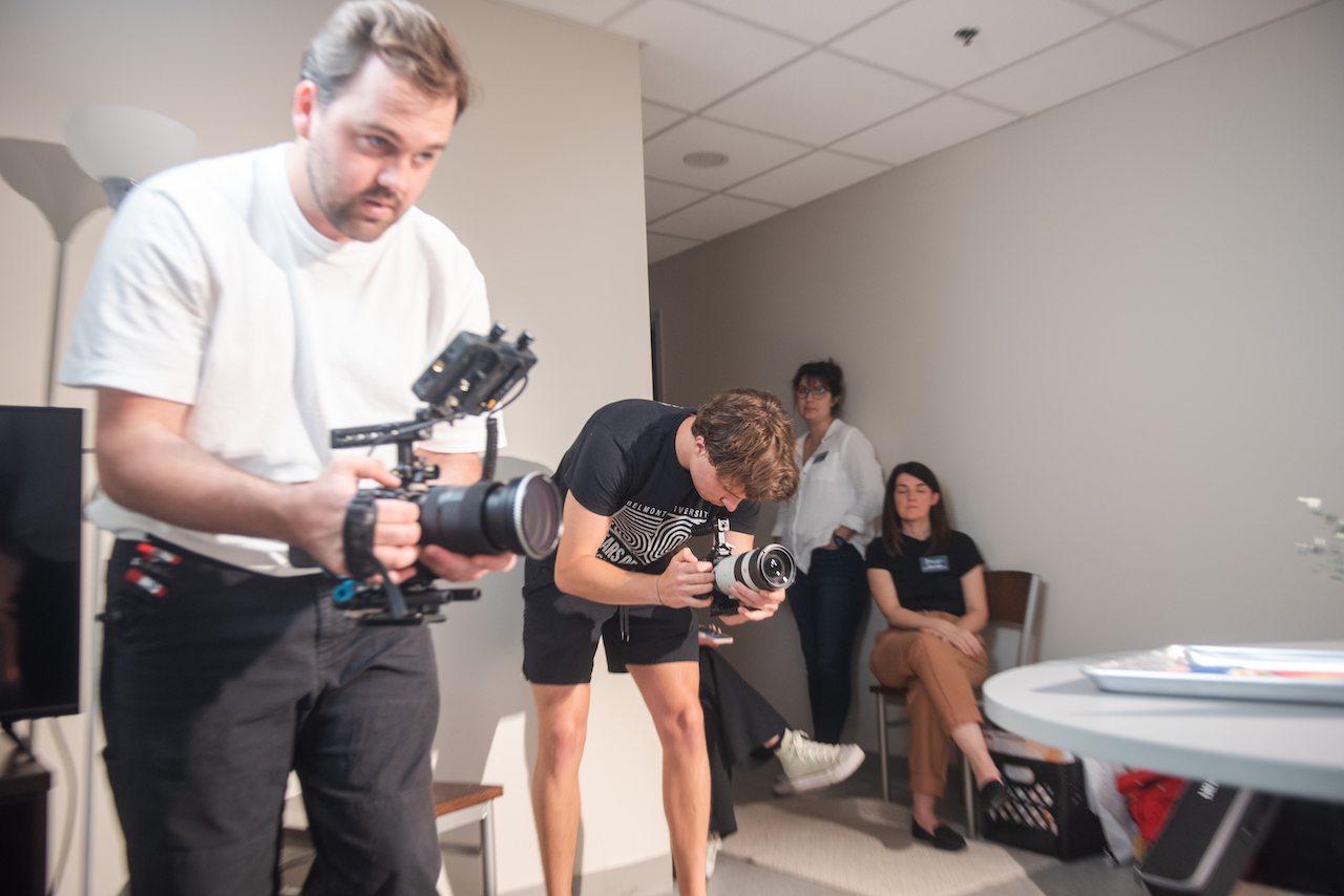 A behind-the-scenes photo of what appears to be a filming or photography session. Two camera operators are working while others observe in what looks like an interior room, possibly documenting the cookie creation process.