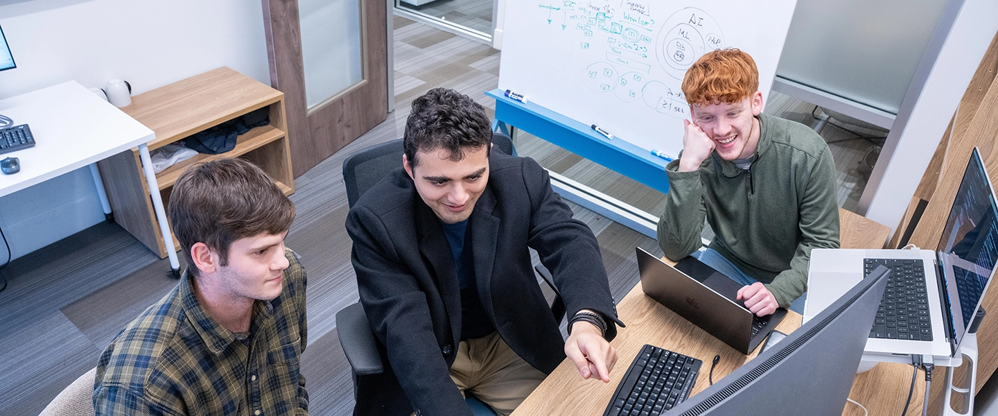 Three male students collaborate at a desk in a modern campus office, reviewing data on a desktop monitor and laptops. A whiteboard with diagrams and notes stands behind them as one student points to the screen while the others look on and smile.
