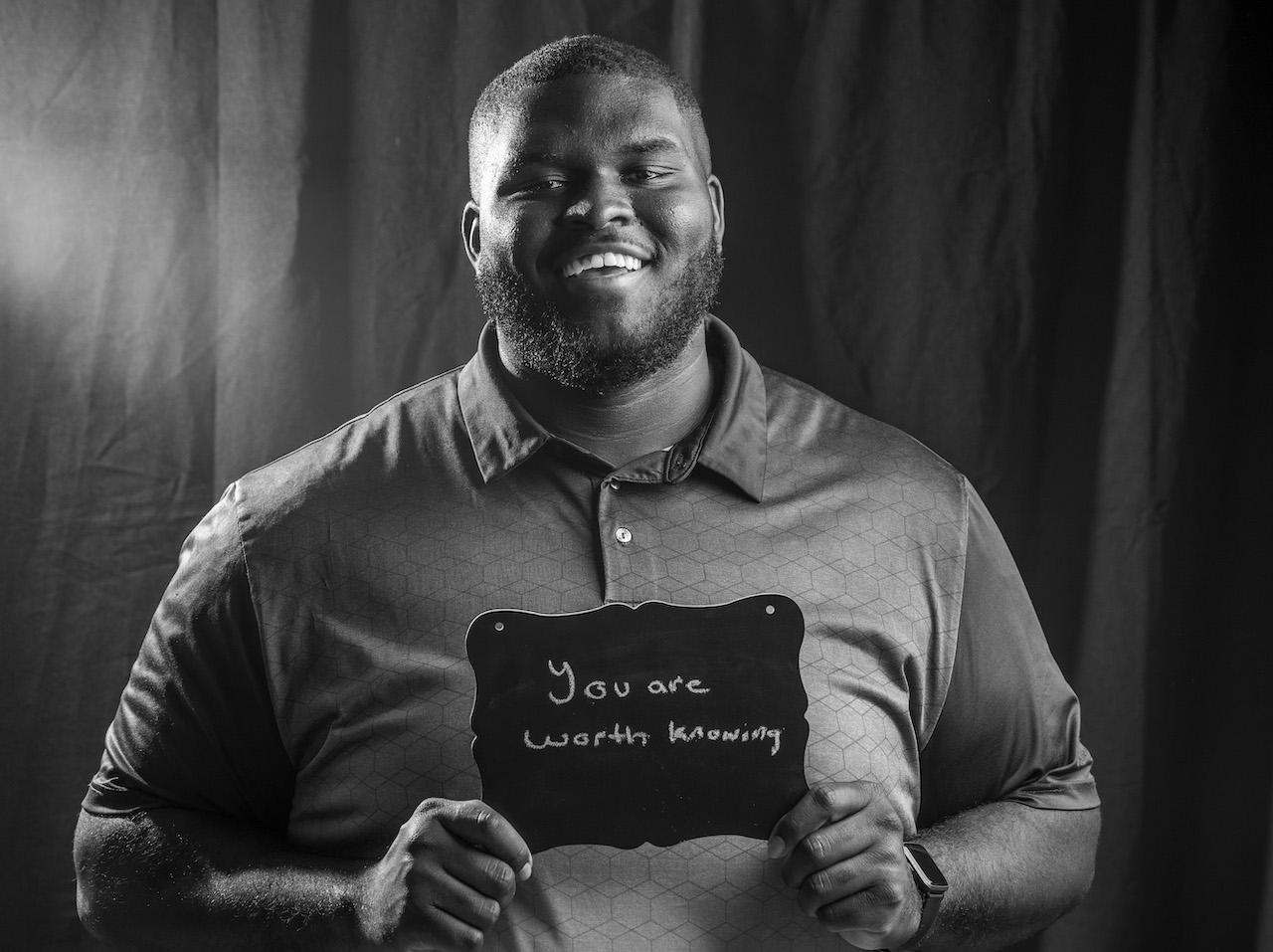A man with a short haircut and beard wearing a collared shirt holds a chalkboard sign reading "You are worth knowing." He has a bright smile and is also wearing a watch.
