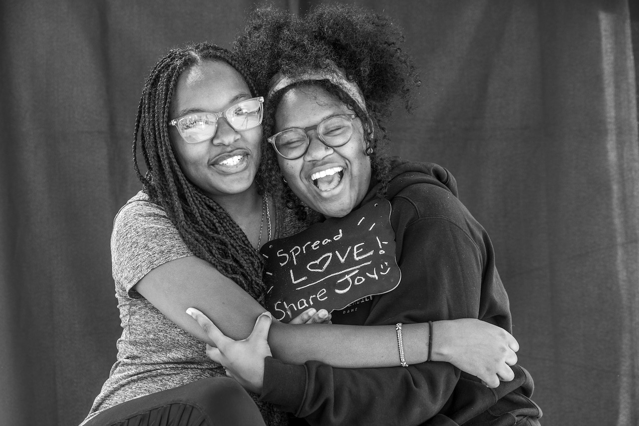 Two young women wearing glasses and embracing closely. Both are laughing joyfully and one holds a small chalkboard sign that reads "Spread LOVE! Share Joy!" They appear to be close friends.