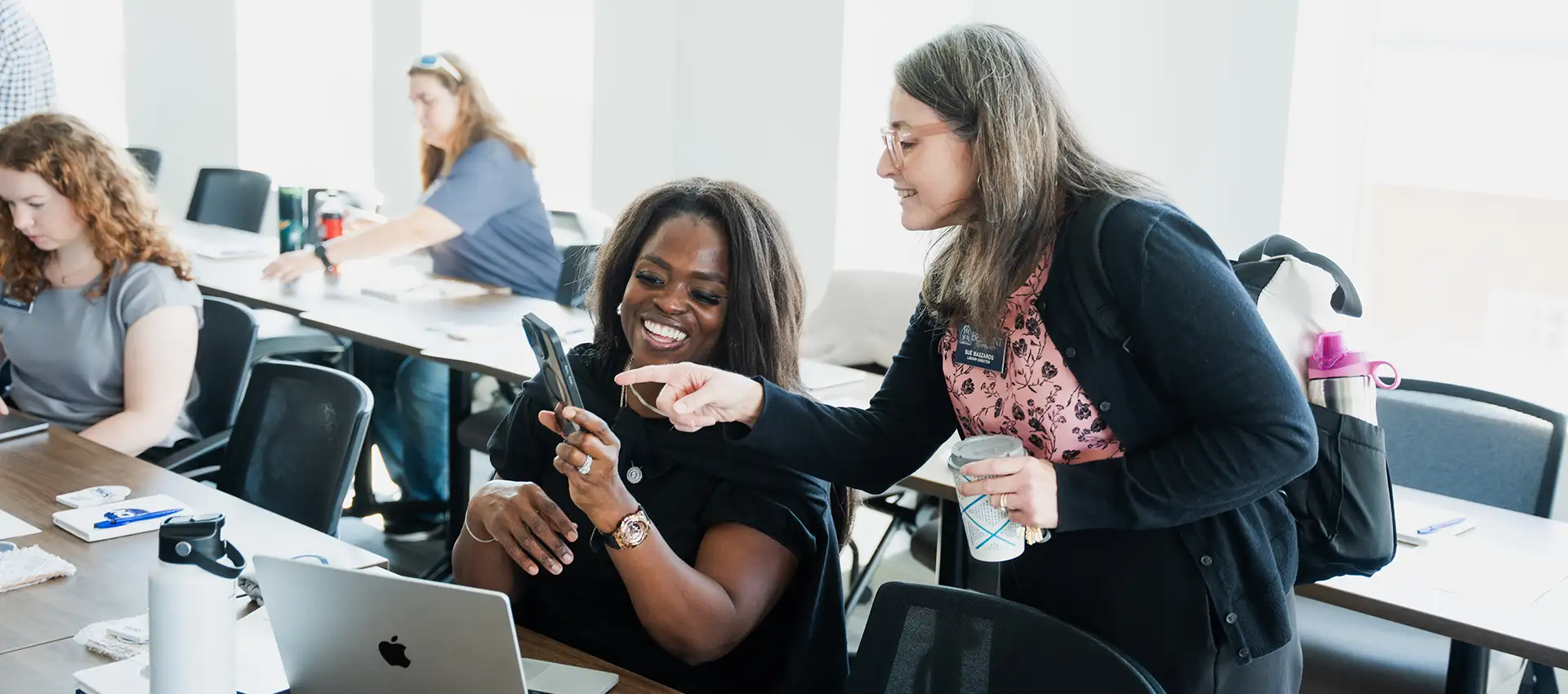 Two women smile and look at a phone together during a classroom workshop, with other participants seated at tables in the background.