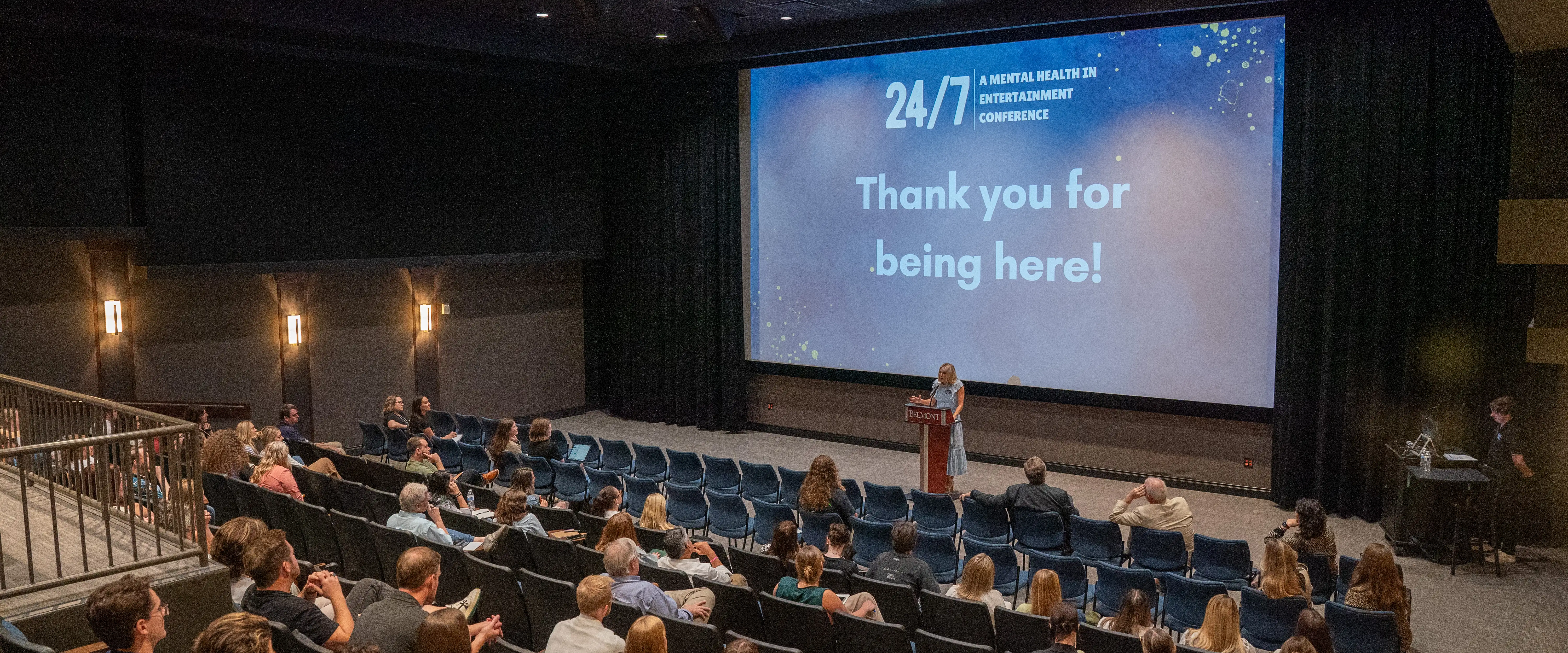 An audience listens as a speaker presents on stage during the 24/7 Mental Health in Entertainment Conference in a theater.