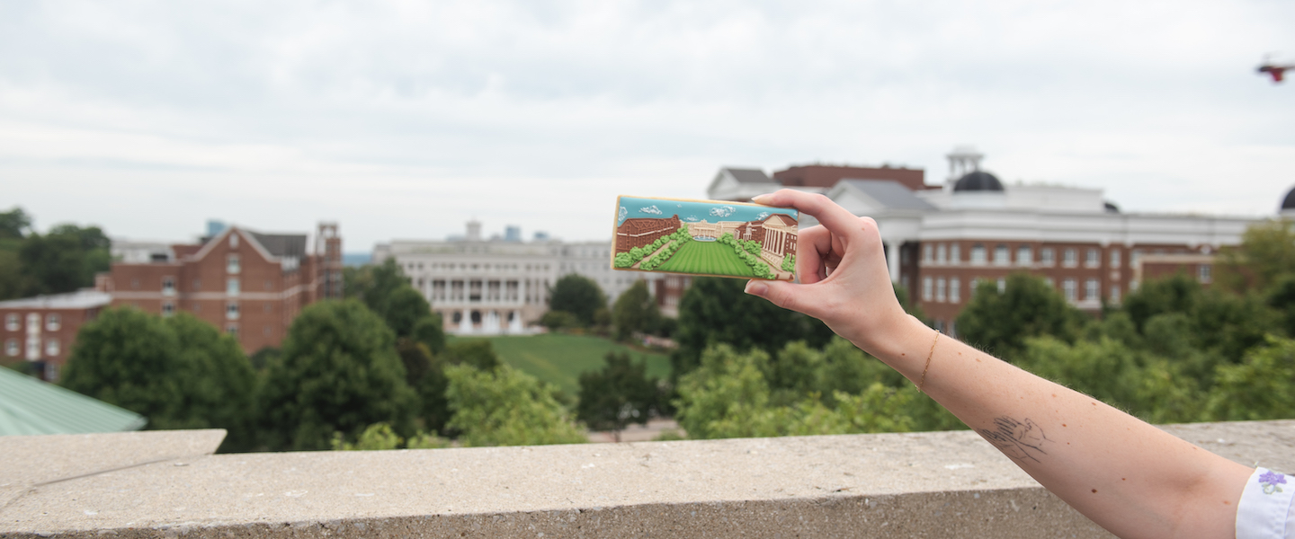 A hand holding up a decorative cookie shaped like Belmont University's campus against the actual campus backdrop. The cookie depicts the green lawn and buildings of the university, creating a visual comparison between the edible art and the real location.