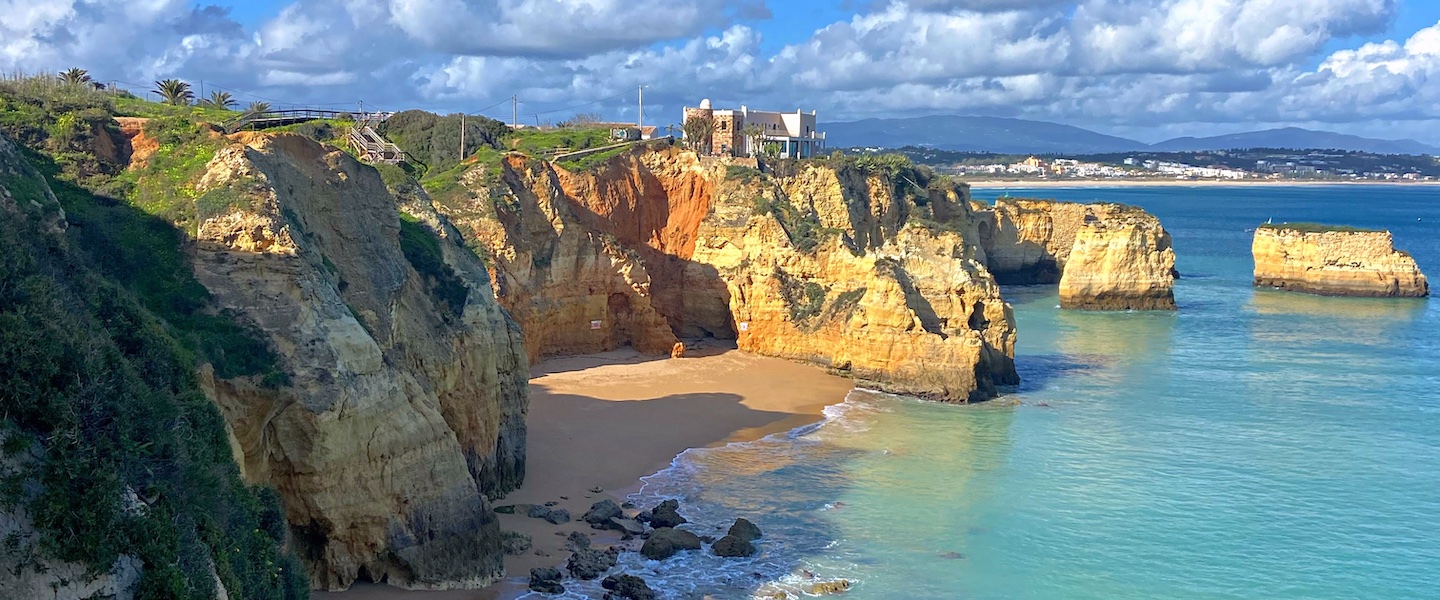 coastal landscape in the Algarve region of Portugal, featuring dramatic golden limestone cliffs surrounding a secluded beach cove. Turquoise waters lap against the sandy shore, with rock formations visible in the ocean. Atop the cliff, buildings and vegetation can be seen against a backdrop of blue sky with fluffy white clouds.