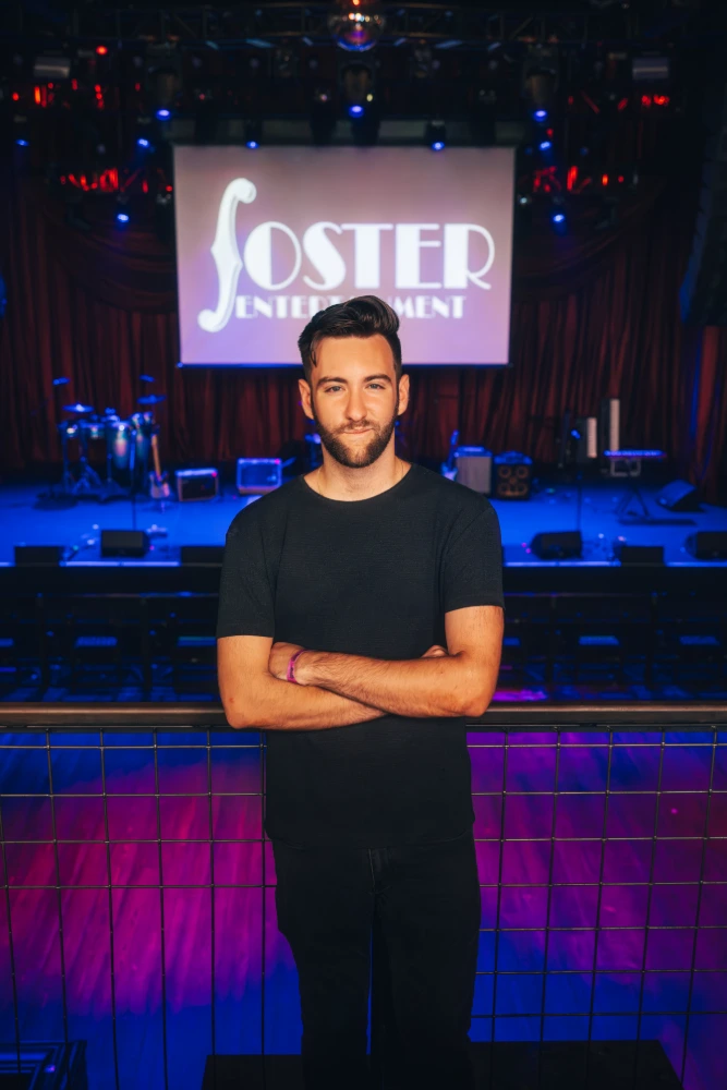 Young man with a beard on stage at a 'Foster Entertainment' event, purple backdrop.
