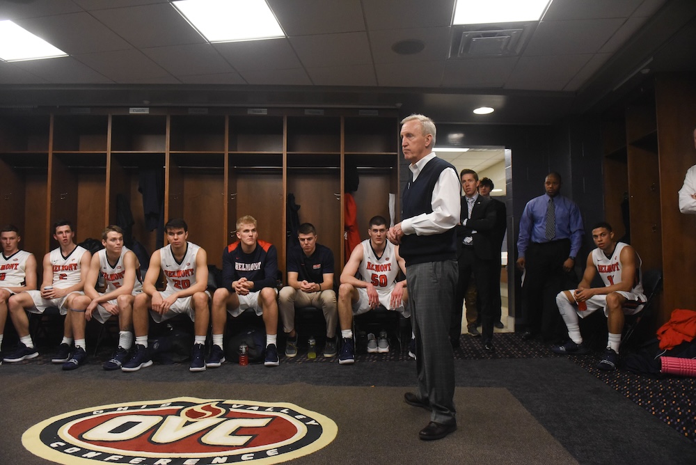A man in a dark vest and gray slacks stands in a locker room addressing a basketball team seated in front of wooden lockers. The players wear white and navy Belmont uniforms, and an OVC (Ohio Valley Conference) logo is on the floor at the bottom of the frame.