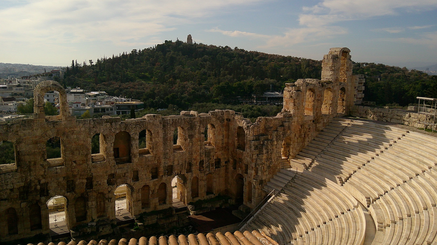 The Odeon of Herodes Atticus, an ancient stone theater on the slopes of the Acropolis in Athens, Greece. The well-preserved semicircular seating faces a stage with the Philopappos Hill visible in the background.