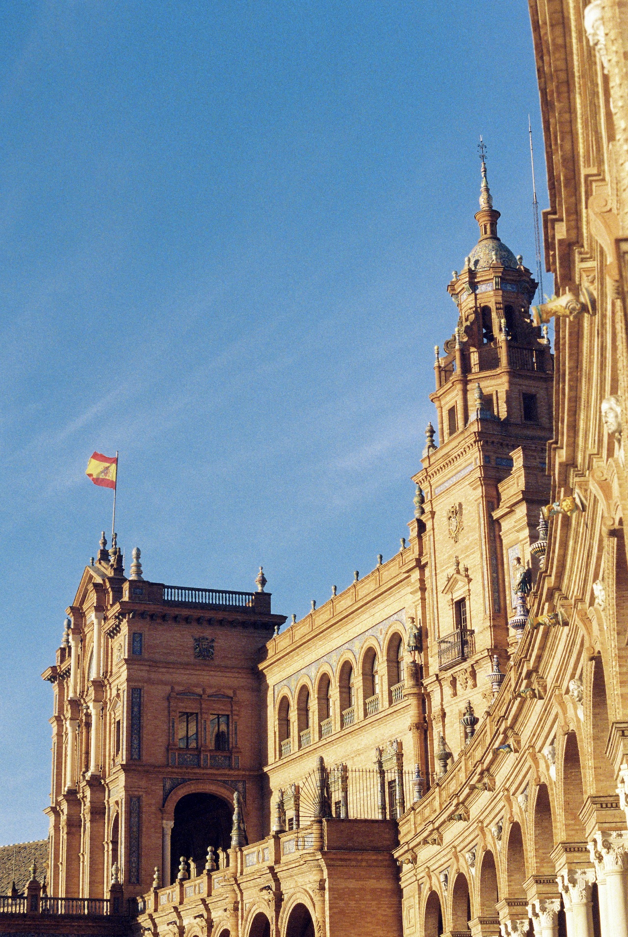 The impressive Plaza de España complex in Seville, Spain, showcasing its Renaissance and Moorish revival architecture bathed in warm sunlight. The Spanish flag flies prominently from one of the towers. The building features ornate stonework, arched galleries, a bell tower with blue-tiled dome, and intricate decorative elements against a clear blue sky.