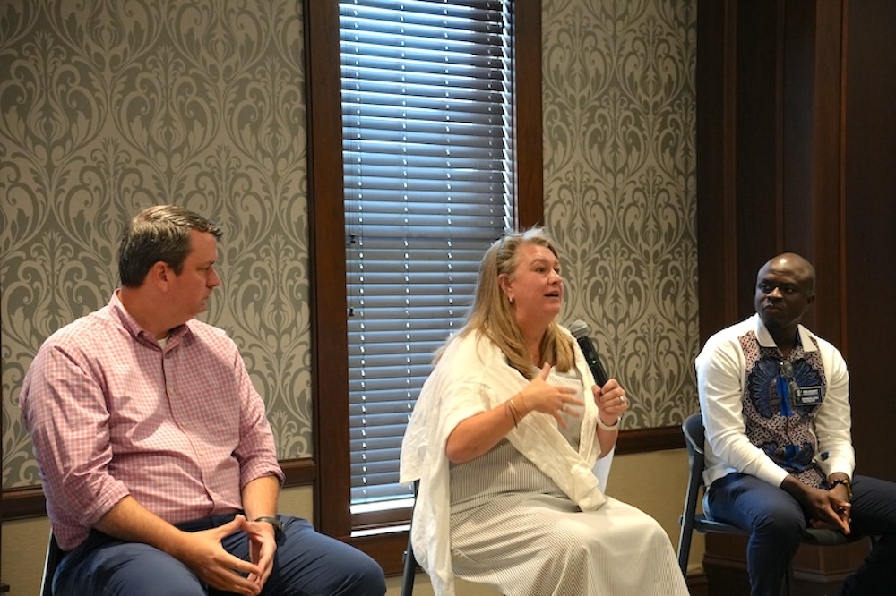 Three people sit in a paneled room with damask wallpaper during a panel discussion. A woman with long blonde hair, wearing a white shawl, speaks into a microphone and gestures with one hand while a man in a pink checkered shirt looks on at left and a man in a patterned shirt with a name badge listens at right.