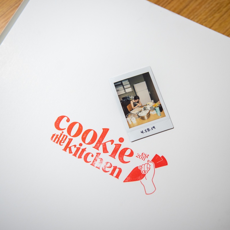 A white box or folder with the "cookie in the kitchen est. 2017" logo in red, featuring an illustration of a hand holding a piping bag. A small polaroid photo dated "4.28.19" is attached, showing someone working in a kitchen.