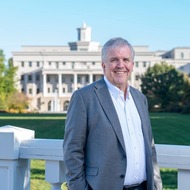 Dr. L. Gregory Jones is standing outdoors, smiling at the camera. He is wearing a light gray suit jacket over a white shirt. In the background, a large building with columns is visible, surrounded by greenery, indicating a bright, clear day. Dr. Jones is the current President of Belmont University.