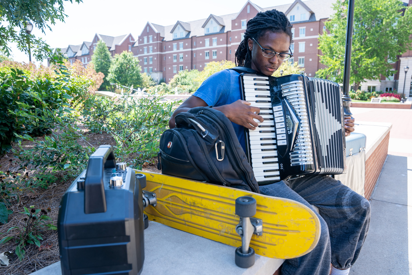 A student with dreadlocks and glasses plays an accordion while seated outdoors on a bench. A yellow skateboard and black backpack rest beside them, with a dormitory-style brick building visible in the background surrounded by trees and landscaping.