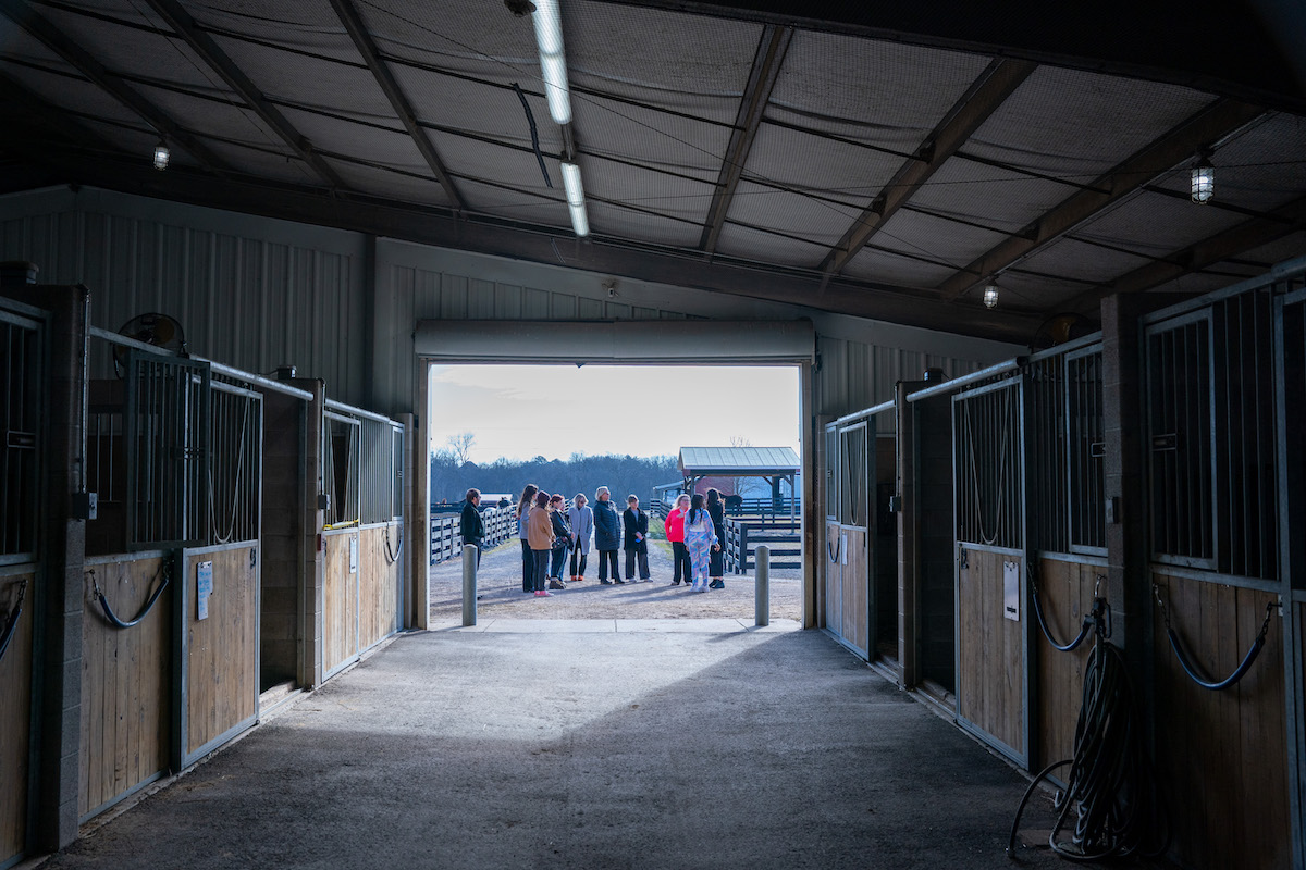 O’More students stand outside the barn at Saddle-Up!, framed by horse stalls and open pasture in the background.