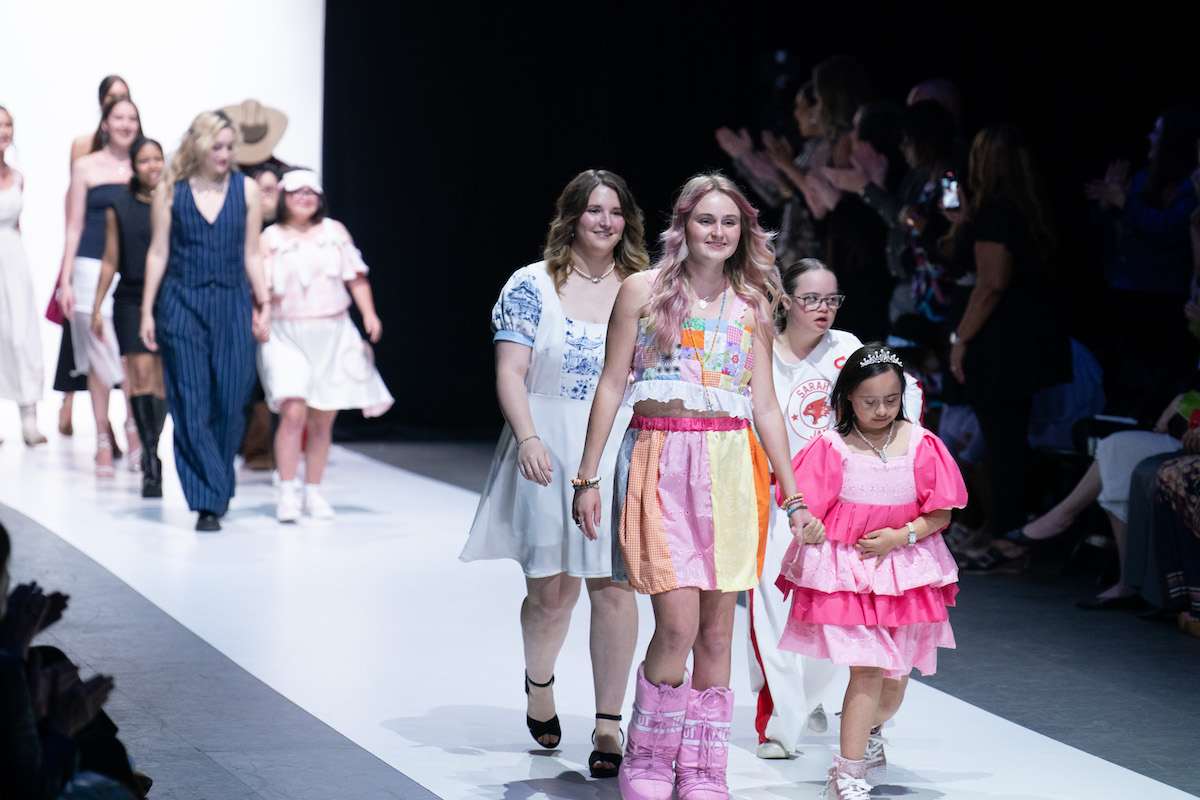 Student designer and young models walk hand-in-hand down the runway, smiling and celebrating a joyful, inclusive fashion show.