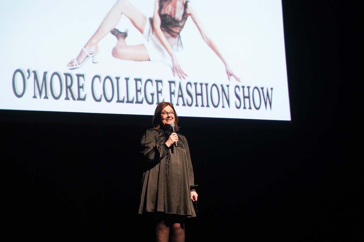 A speaker in a black dress stands on stage under the projected O’More Fashion Show title, addressing the audience with a smile.