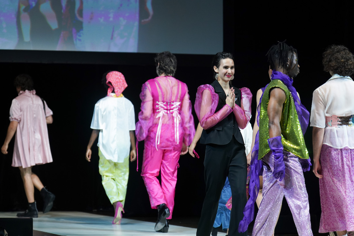 A student designer in bright pink sleeves takes a bow alongside models in glittering and sheer avant-garde looks.