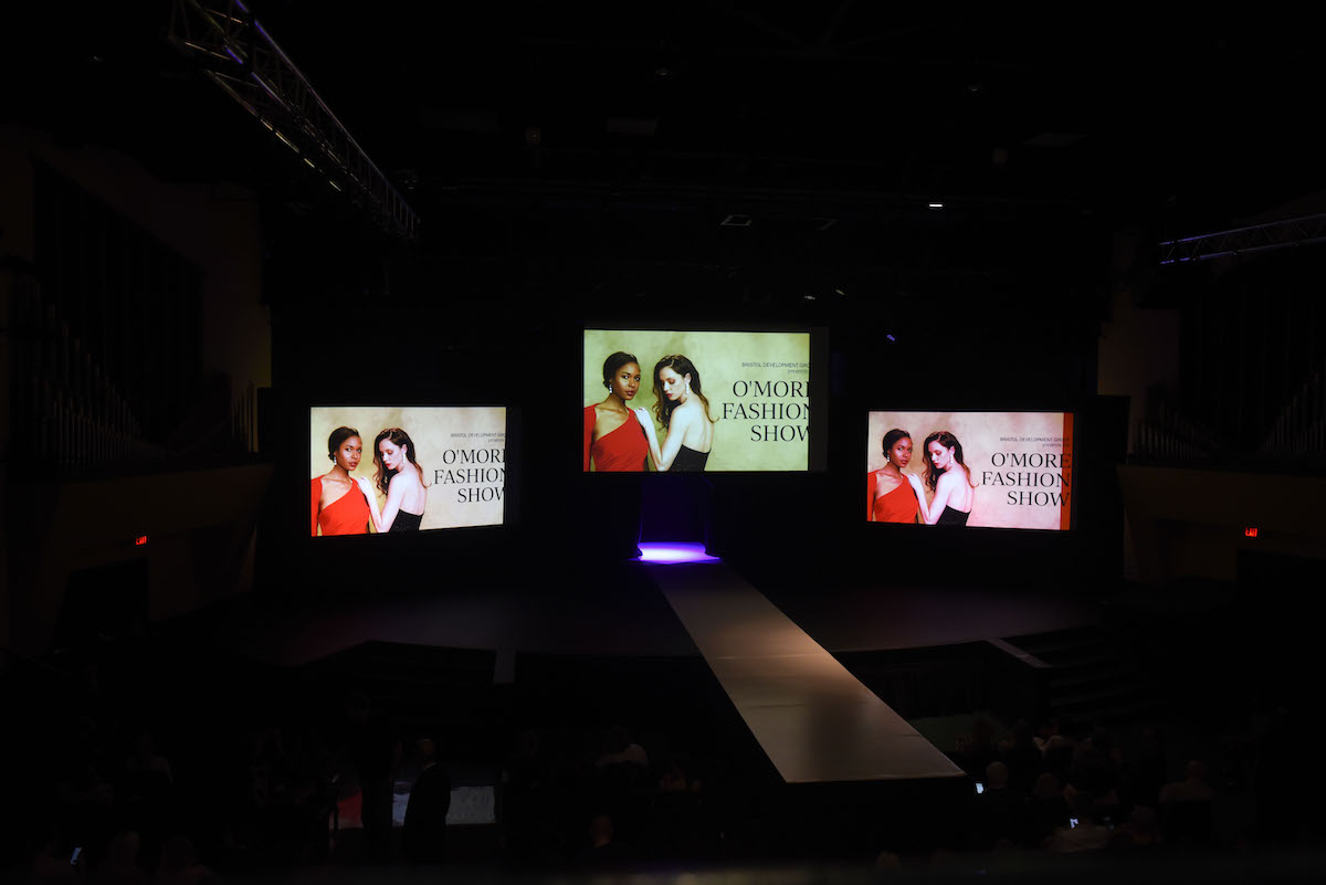 A wide runway flanked by screens displays the O’More Fashion Show title card featuring two student models in red and black gowns.