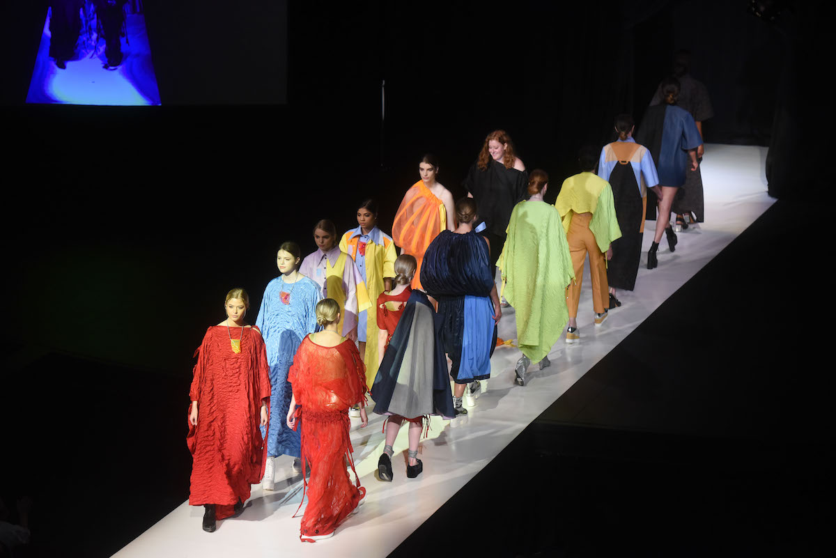 A lineup of models in vibrant, sculptural garments—red, blue, orange, lime, and gold — circles the runway.