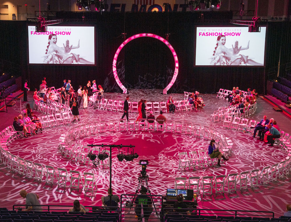 Overhead view of the 2025 O’More College Fashion Show, featuring a glowing figure-eight runway with white chairs arranged around it in a large indoor arena, illuminated with pink lighting and decorative projections.