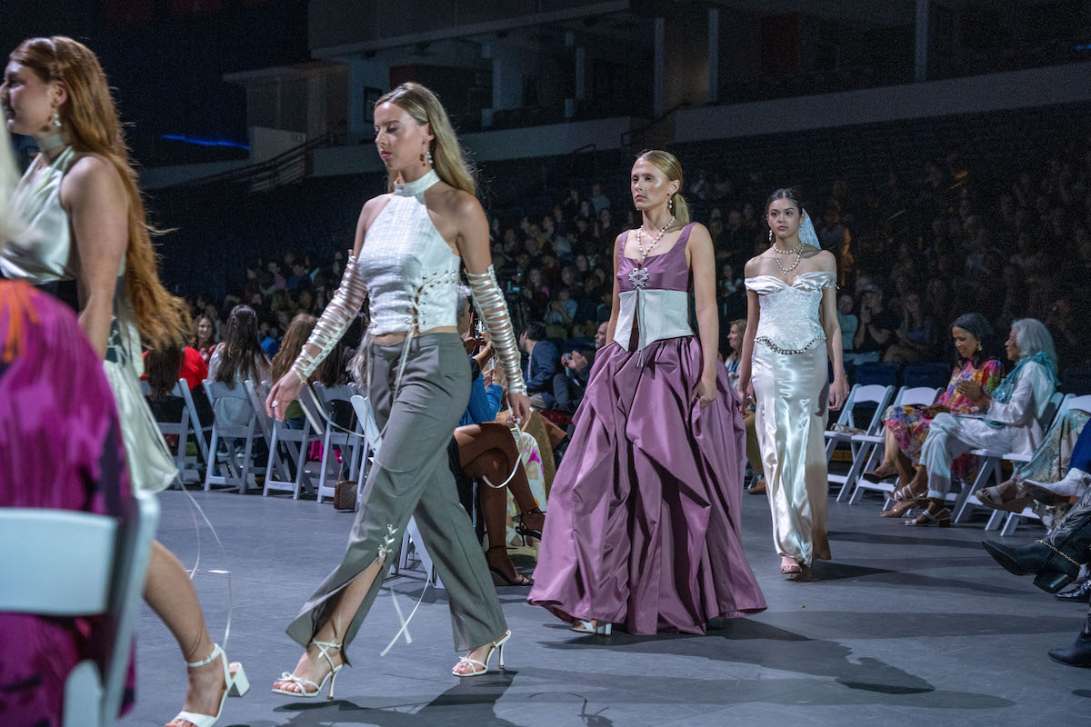 A trio of models showcases structured, high-fashion eveningwear featuring metallic finishes, pearl accents, and dramatic silhouettes in white, lavender, and deep plum on the runway.