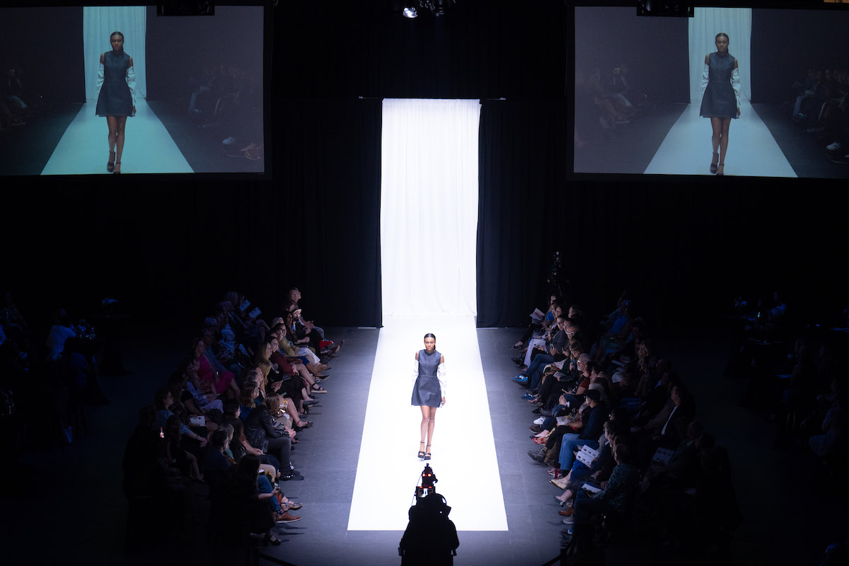 A solo model walks a stark white runway surrounded by audience seating, with her image projected large on dual screens above.