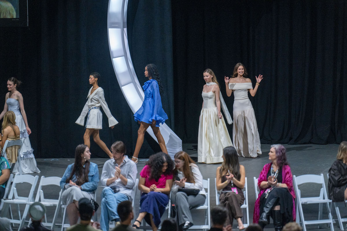 Models in cream and ivory gowns, including a striking blue mini-dress, walk beneath a glowing arch during the O’More Fashion Show.