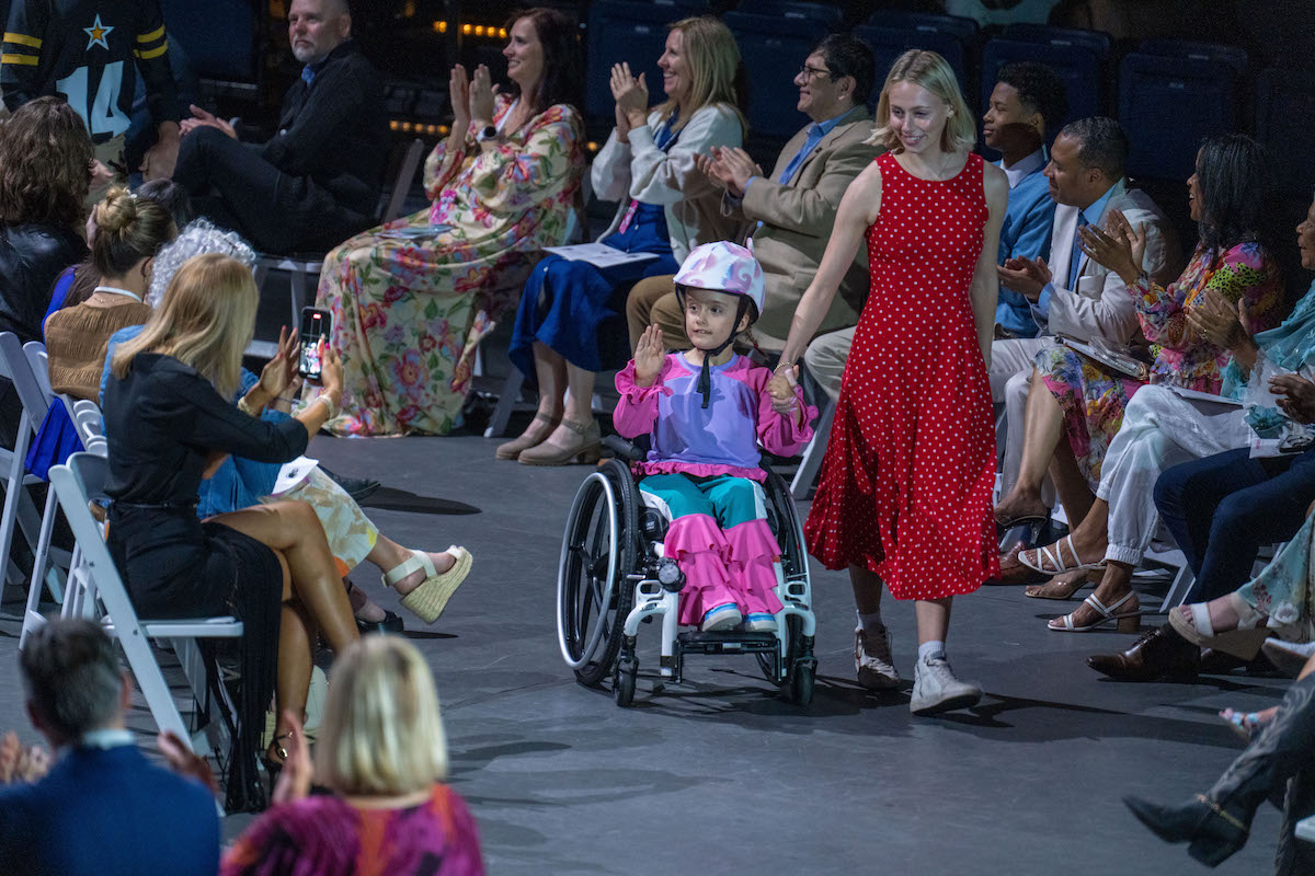 Young model in wheelchair and pink outfit is escorted down the runway by a designer in red dress.