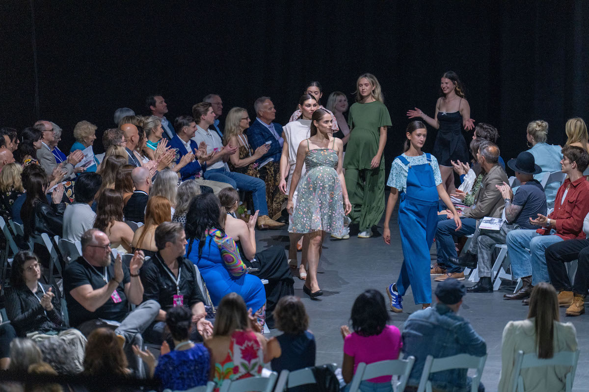 Models walk figure-eight runway in casual and maternity wear as audience watches intently.