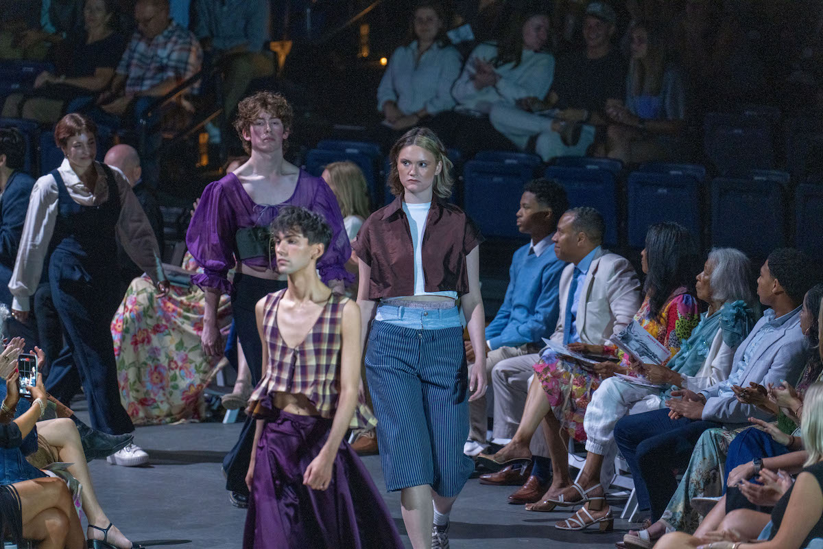Models display bold, gender-fluid fashion with dramatic silhouettes and dark tones during the runway show.