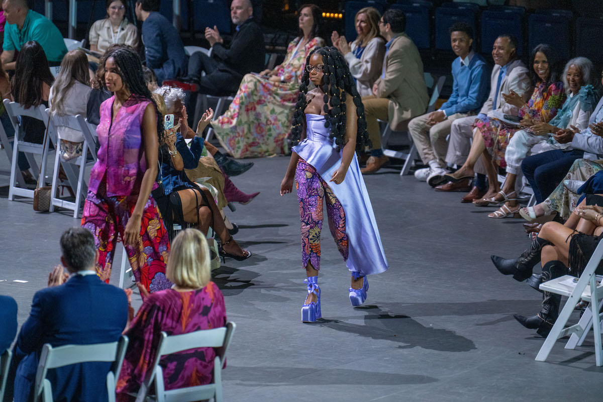 Models showcase colorful, patterned garments—one in flowing lavender asymmetrical top, the other in sheer pink ensemble.