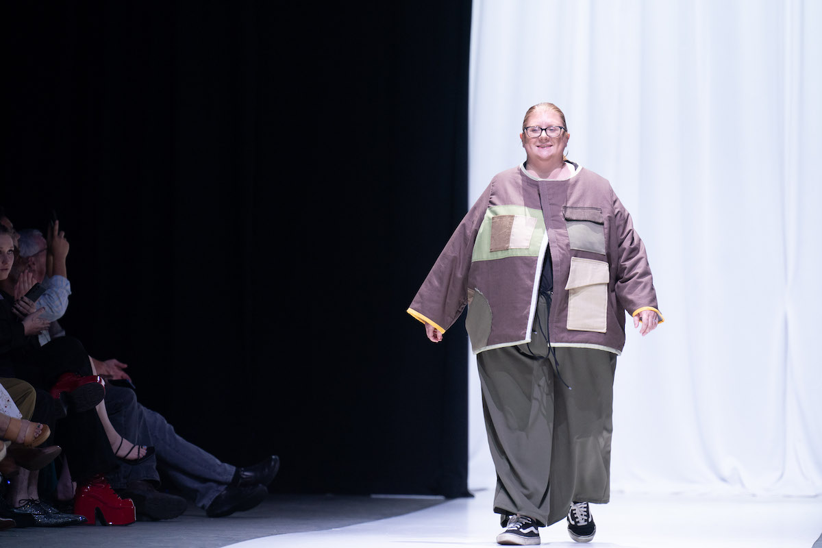 A smiling designer in a brown and green jacket and wide-leg pants walks the runway.