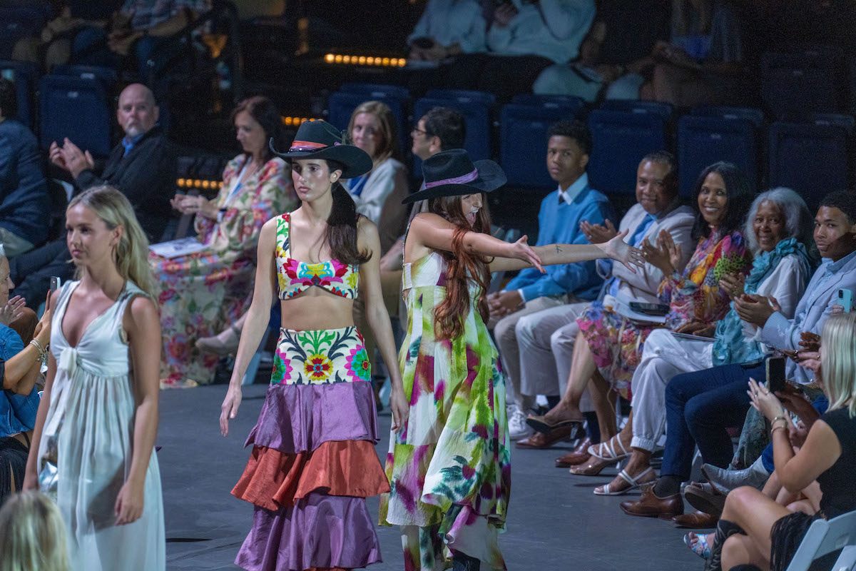 Models in colorful, Western-inspired outfits—including tiered skirts, a floral crop top, and wide-brim hats—energize the runway with playful gestures and bold prints.