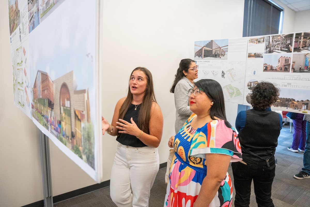 Student discusses project details with a guest in front of a design board featuring colorful building renderings.